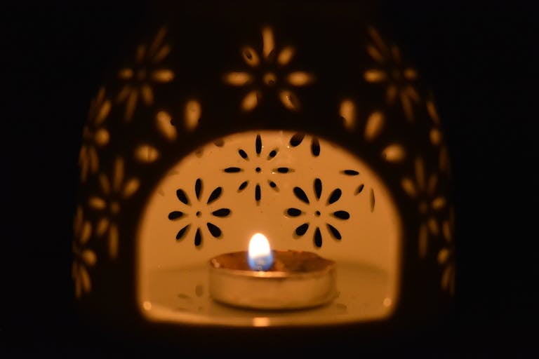 A close-up of a decorative ceramic candle holder with a burning tealight inside, creating a warm floral pattern.
