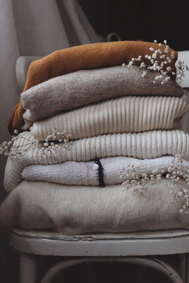 Aesthetic stack of beige and brown woolen sweaters on a chair indoors, adorned with dried flowers.