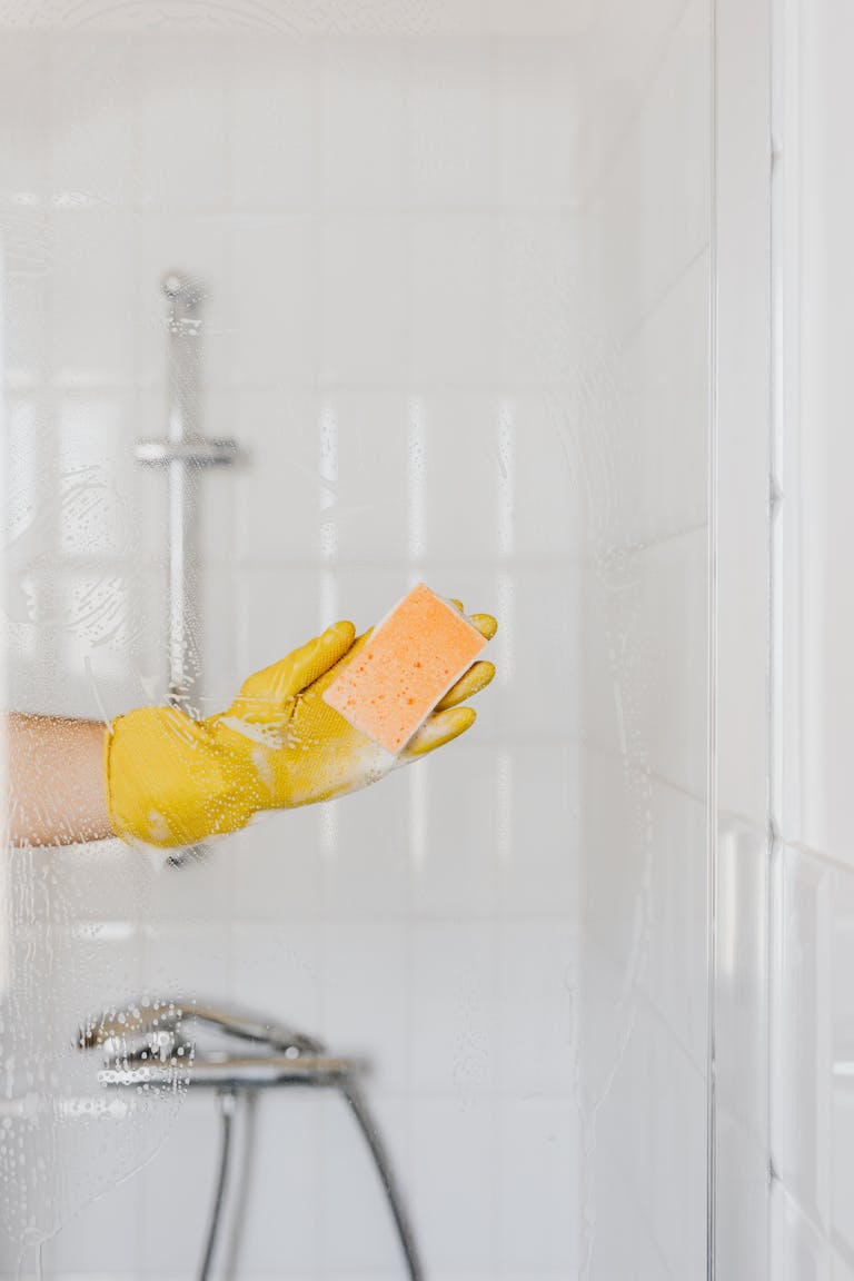 Crop anonymous person in rubber protective glove washing shower cabin door with sponge and detergent