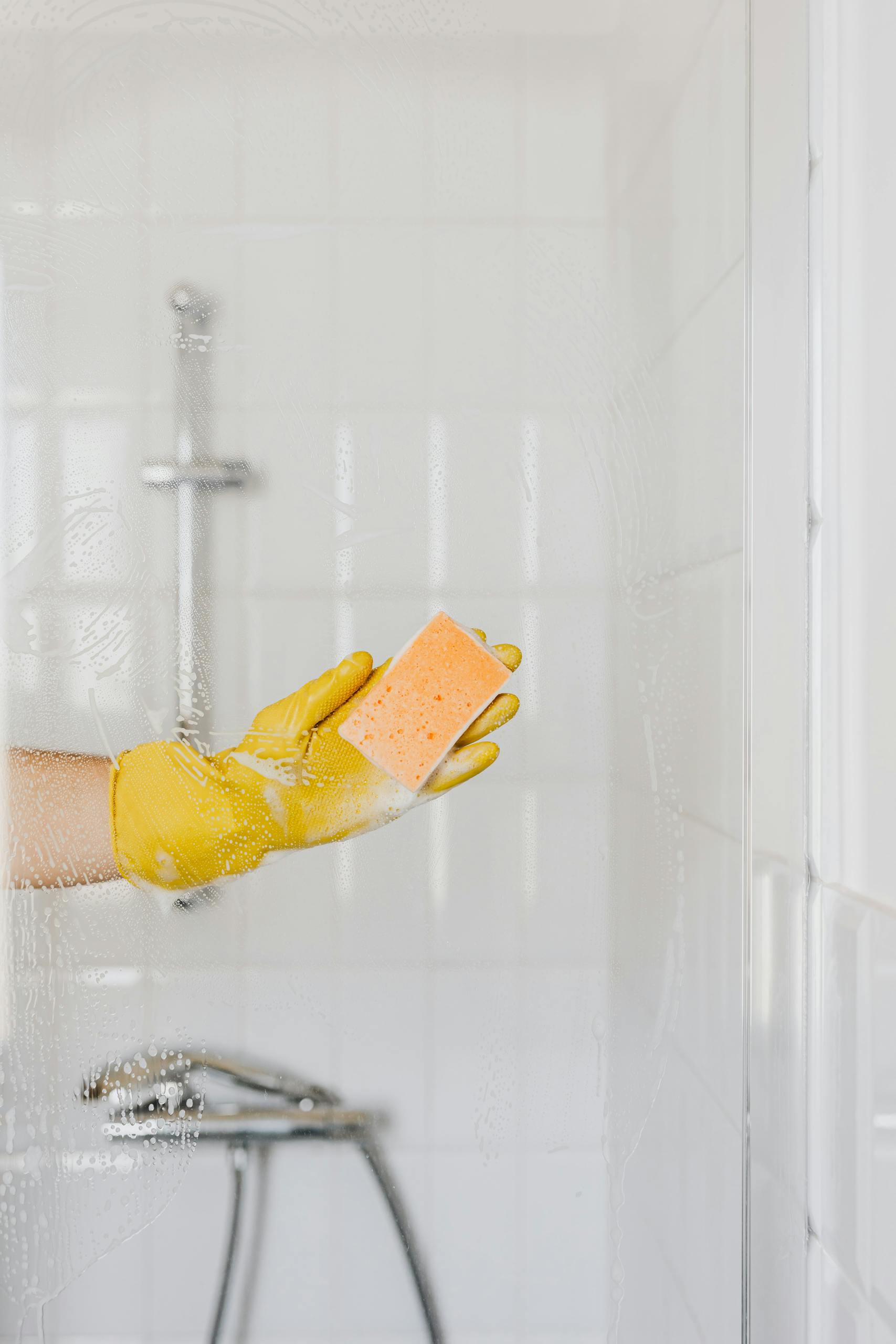 Crop anonymous person in rubber protective glove washing shower cabin door with sponge and detergent
