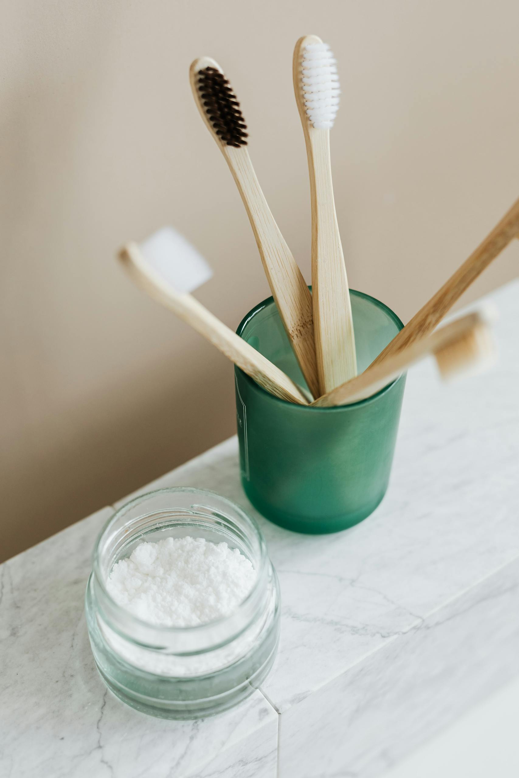 From above of assorted bamboo eco friendly toothbrushes in translucent green holder near opened glass reusable jar with white toothpaste arranged on marble table against beige wall