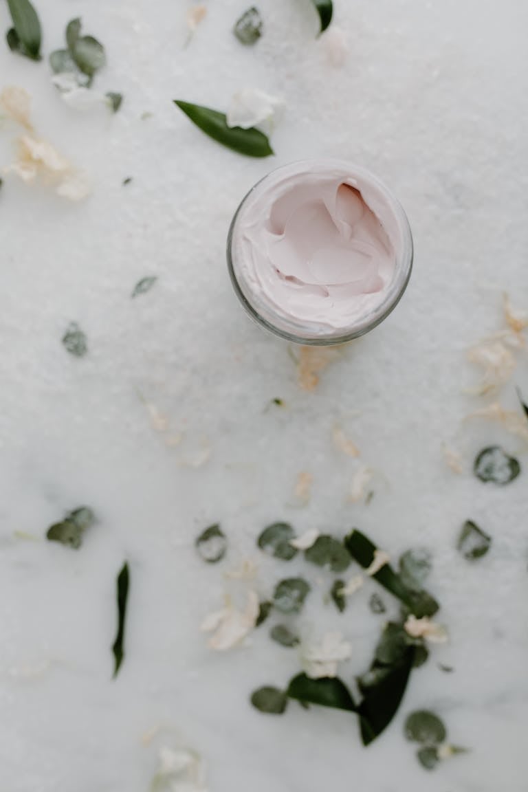 Top view of an open cream jar surrounded by green leaves and white flowers on a white surface.