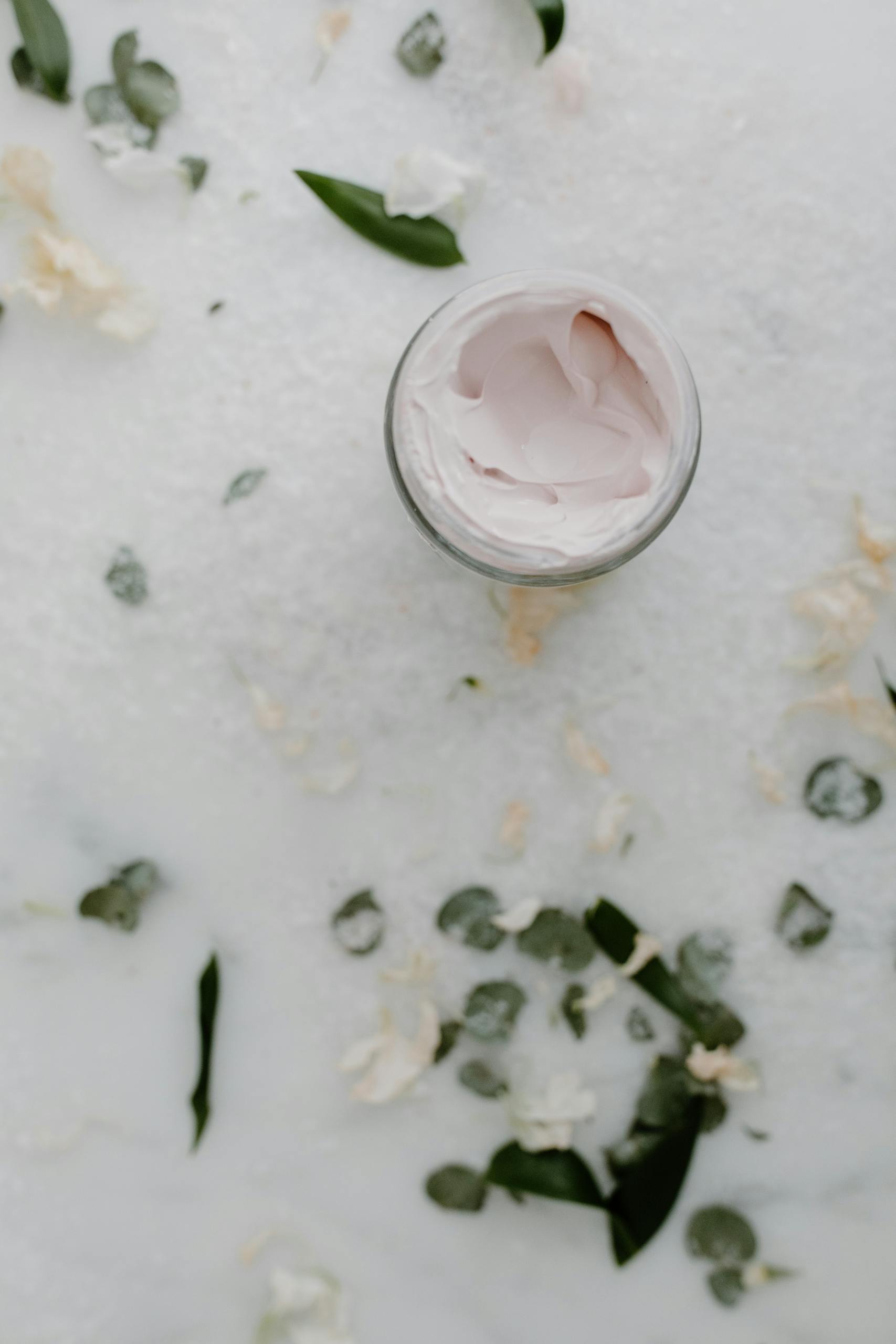 Top view of an open cream jar surrounded by green leaves and white flowers on a white surface.